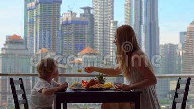 年轻的女家庭教师和一个男孩在阳台上吃早餐，俯瞰市中心的摩天大楼